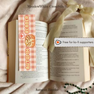 May include: A cross-stitch bookmark with the words "AUTUMN READER" and a pumpkin design, placed in an open book. The bookmark has a checkered pattern in orange and white. The text "Autumn Reader" is at the bottom.