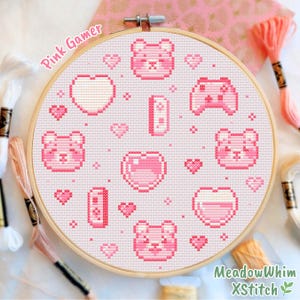 May include: A pink cross-stitch embroidery hoop with a gamer theme. The design features pixelated hearts, bears, and game controllers. The text "Pink Gamer" and "MeadowWhim XStitch" are visible. Embroidery floss in shades of pink and white surrounds the hoop.