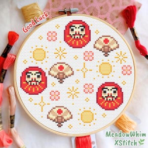 Könnte beinhalten: Ein Kreuzstich-Stickrahmen mit einem Daruma-Puppen-Design, Sonnen und floralen Elementen in Rot, Gelb und Braun. Der Text "Good Luck" ist sichtbar. Stickgarn umgibt den Rahmen.