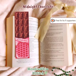 Op de afbeelding: Een handgemaakt bladwijzer met een kruissteekontwerp van een chocoladereep en een roze snoepreep met hartjes. De bladwijzer is in een open boek geplaatst. De tekst op de bladwijzer luidt "Midnight Chocolate" en "MeadowWhim XStitch".