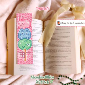 Może przedstawiać: Zakładka do książki z haftem krzyżykowym przedstawiająca różowego królika, niebieskiego kota i zielone kulki, z japońskim słowem "Dango" i tekstem "MeadowWhim XStitch". Zakładka znajduje się w otwartej książce.