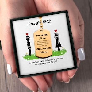 Può includere: Un ciondolo a forma di targhetta dorata con la scritta "Proverbi 18:22" e una citazione sul trovare una moglie. Il ciondolo è esposto in una scatola nera con sfondo bianco e illustrazioni.