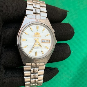 May include: A silver Orient wristwatch with a white face, gold detailing, and a day-date display. The watch has a silver metal strap and is shown against a black background. The watch face features the Orient logo and the text "Crystal 21 Jewels".
