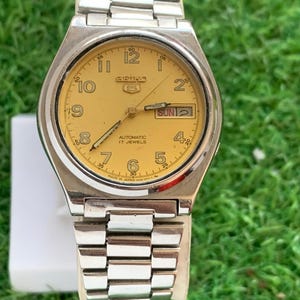 May include: A vintage Seiko automatic wristwatch with a silver-tone metal band and case. The watch face is a light yellow color with black numerals and hands. The day and date are displayed at the 3 o'clock position. The watch is displayed on a white surface.