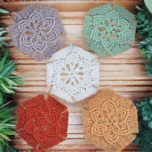 May include: Five handmade macrame coasters in various colors: gray, green, white, rust, and tan. Each coaster features a floral design with a fringed edge. The coasters are arranged on a wooden surface with greenery.