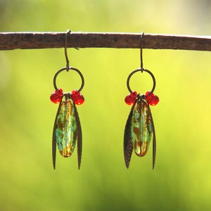 May include: A pair of dangle earrings featuring a central elongated green and amber glass bead flanked by two dark metal leaf-shaped components. Each earring is topped with a small bronze-toned ring and two vibrant red glass beads.