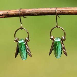 May include: A pair of brass-toned earrings with a green, teardrop-shaped bead and bronze-colored wing accents. The earrings are suspended from a thin, curved hook. The background is a soft, blurred green.