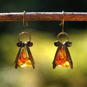 May include: A pair of handcrafted earrings with a unique design. Each earring features a gold-colored hook, a small ring, and a cluster of dark beads. Below, a cage-like structure holds amber-colored, faceted glass pieces, creating a warm, glowing effect. Jewelry.