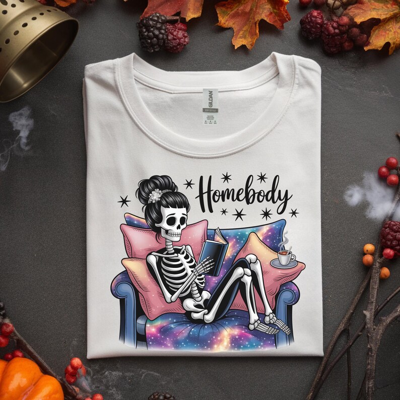 Op de afbeelding: Wit T-shirt met een illustratie van een skelet dat een boek leest op een bank. Het skelet heeft een knot en is omringd door kussens en een kopje koffie. Op het shirt staat het woord "Homebody".