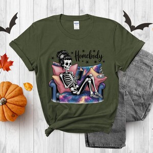 Op de afbeelding: Olijfgroen t-shirt met een skelet dat een boek leest op een bank. Het skelet is omringd door kussens en een kopje koffie. De tekst "Homebody" staat boven het skelet. Halloween thema.