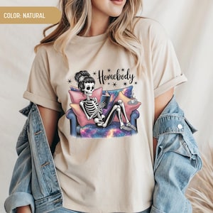 Op de afbeelding: Beige t-shirt met een skelet dat een boek leest op een bank. Het skelet heeft een bloemenhaarstuk. De bank heeft een galaxy-ontwerp. De tekst "Homebody" staat boven het skelet. Het shirt is neutraal van kleur.