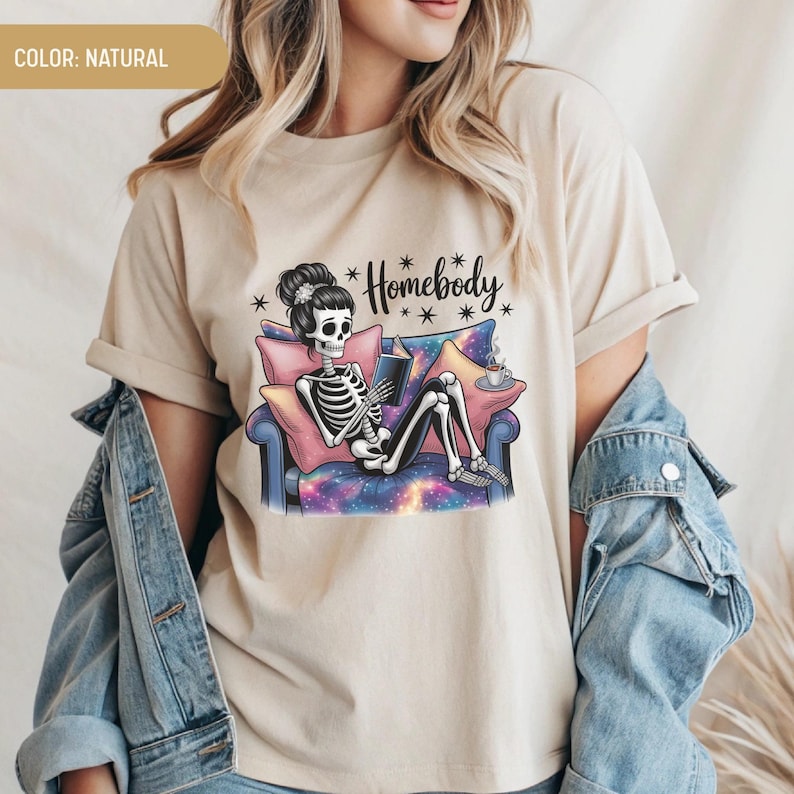 Op de afbeelding: Beige t-shirt met een skelet dat een boek leest op een bank. Het skelet heeft een bloemenhaarstuk. De bank heeft een galaxy-ontwerp. De tekst "Homebody" staat boven het skelet. Het shirt is neutraal van kleur.