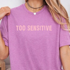 May include: A heathered purple short-sleeve t-shirt featuring the text "TOO SENSITIVE" printed in a light peach color across the chest. The shirt has a crew neckline and a standard fit, showcasing a simple design with clear, bold lettering.