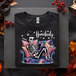 Op de afbeelding: Zwart T-shirt met een illustratie van een skelet dat een boek leest op een bank. Het skelet heeft een knot en het shirt heeft het woord "Homebody". De bank heeft een galaxy print.