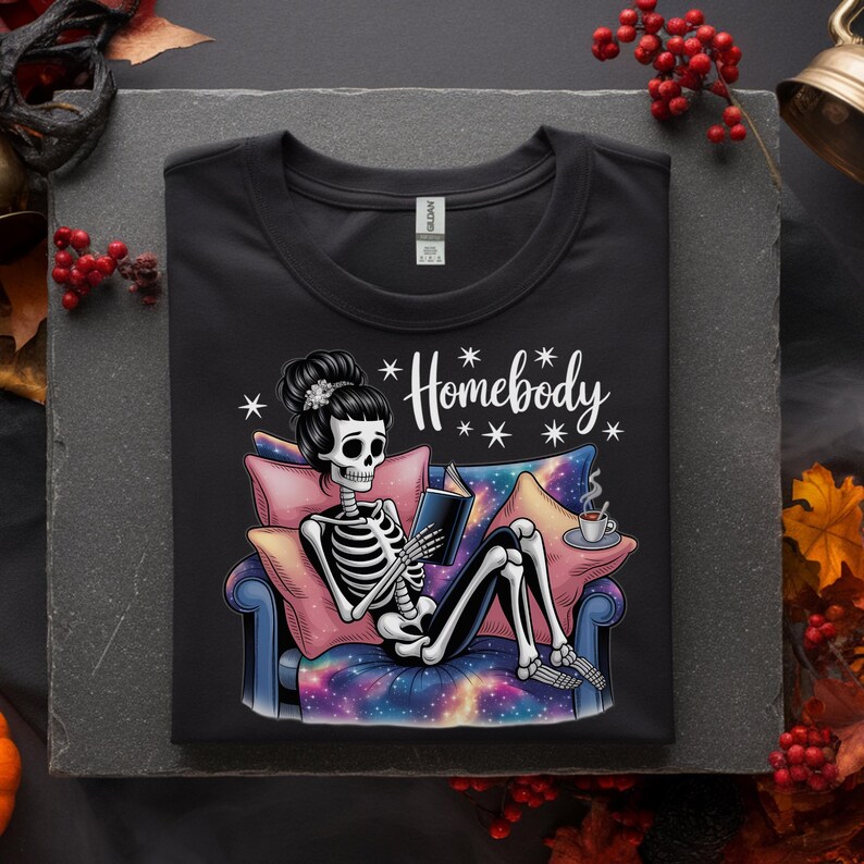 Op de afbeelding: Zwart T-shirt met een illustratie van een skelet dat een boek leest op een bank. Het skelet heeft een knot en het shirt heeft het woord "Homebody". De bank heeft een galaxy print.