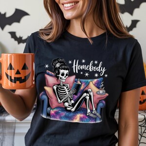 Op de afbeelding: Zwart T-shirt met een skelet dat een boek leest op een bank. Het skelet heeft een bloemenkapsel. Op het shirt staat "Homebody" in witte letters. Een oranje pompoenmok wordt in de hand gehouden. Halloween-thema.