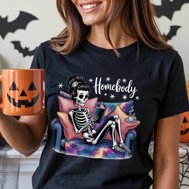 Op de afbeelding: Zwart T-shirt met een skelet dat een boek leest op een bank. Het skelet heeft een bloemenkapsel. Op het shirt staat "Homebody" in witte letters. Een oranje pompoenmok wordt in de hand gehouden. Halloween-thema.