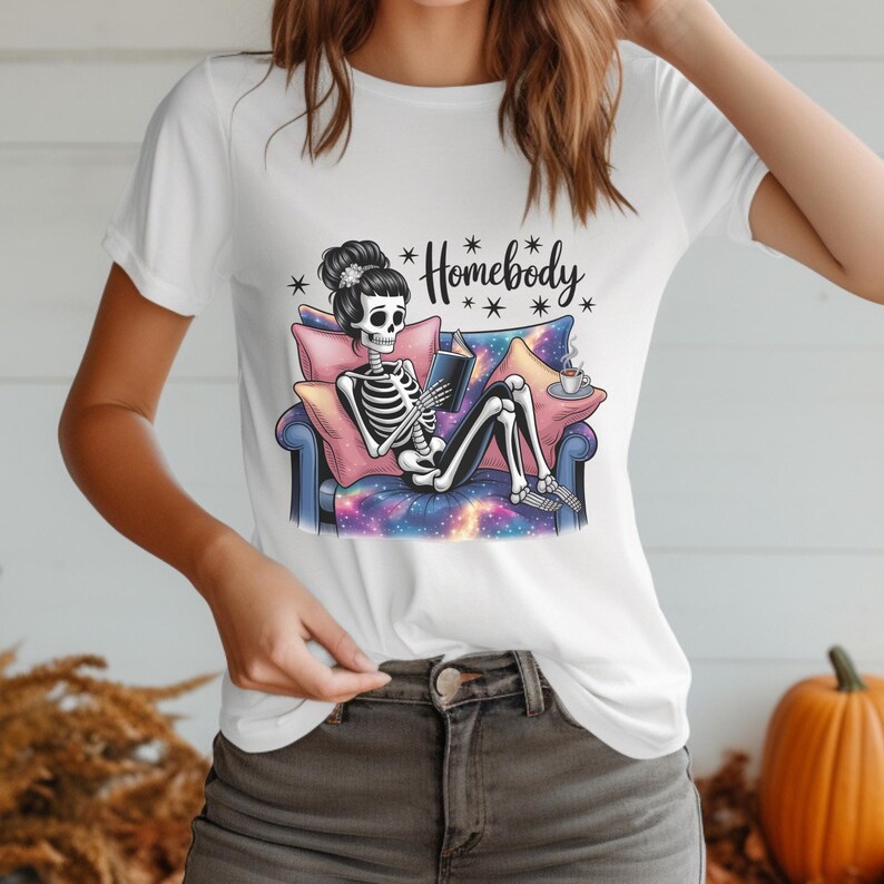 Op de afbeelding: Wit T-shirt met een skelet dat ontspant op een bank, een boek leest en koffie drinkt. De bank heeft een galaxy-thema. De tekst "Homebody" staat boven het skelet. Het ontwerp is in zwart, wit, roze, blauw en paars.