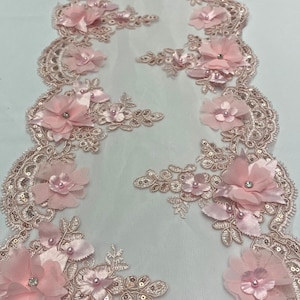 May include: Pink floral lace trim with 3D fabric flowers, sequins, and pearl accents. The trim features a scalloped edge and is ideal for embellishing garments, accessories, or craft projects. The delicate design is perfect for adding a touch of elegance.