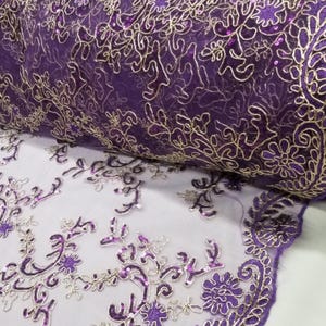 May include: A roll of purple fabric with gold embroidered floral patterns and sequins. The fabric has a delicate, sheer appearance, with intricate detailing in the embroidery. The design features swirling lines and floral motifs, creating a luxurious look.