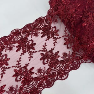 May include: Burgundy lace fabric with floral and leaf patterns. The fabric is embellished with sequins and has a scalloped edge. The lace is rolled up, showing the intricate details and texture of the design.