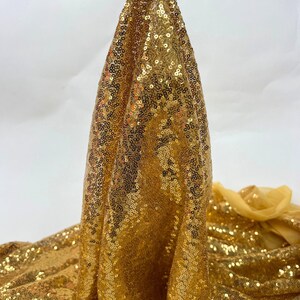 May include: A close-up of a gold sequin fabric. The fabric is held up, showcasing its draped texture and the way the sequins catch the light. The sequins are densely packed, creating a shimmering effect. The background is plain white.
