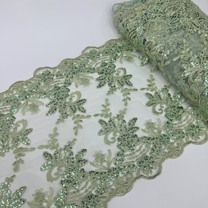 May include: Sage green lace fabric with floral and leaf patterns, embellished with sequins. The fabric is rolled up and spread out, showcasing the intricate design and scalloped edges. The lace is on a white background.