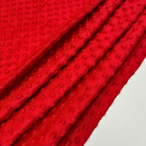 May include: A close-up of a folded, bright red fabric with a textured, dotted pattern. The fabric appears soft and plush, with visible folds and shadows that add depth. The material is likely a blanket or throw.