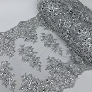 May include: A roll of silver lace fabric with intricate floral embroidery and sparkling sequins. The fabric is a light gray color and has a delicate, sheer appearance. The edge of the fabric has a scalloped design.