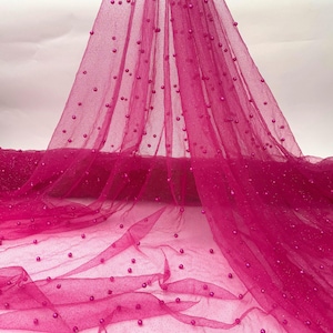 May include: A vibrant fuchsia tulle fabric adorned with scattered, small, round beads. The sheer material is draped, creating soft folds and a delicate texture. The beads add a touch of sparkle and dimension to the fabric, suitable for various craft projects.