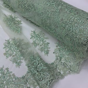 May include: A roll of mint green lace fabric with intricate floral patterns and shimmering sequins. The delicate lace features detailed embroidery and a scalloped edge, perfect for elegant apparel or craft projects.