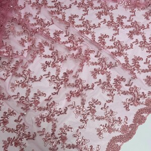 May include: Close-up of a sheer, dusty rose lace fabric with intricate floral embroidery and scattered sequins. The delicate design features detailed patterns and a scalloped edge, perfect for elegant apparel or craft projects.