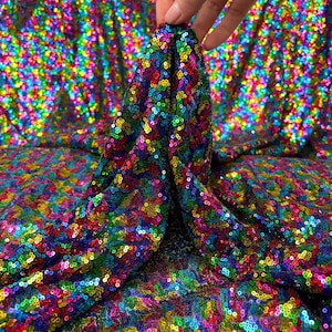 May include: A vibrant fabric covered in small, colorful sequins. The sequins are in a rainbow of colors, including red, orange, yellow, green, blue, and purple. The fabric is being held up, showing its texture and drape.