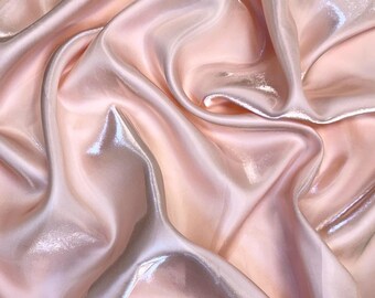 May include: A close-up shot of a flowing, iridescent fabric in shades of pale pink and peach. The material is draped and folded, creating soft curves and highlights. The fabric has a subtle sheen, reflecting light and adding depth to the image.