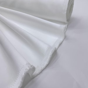 May include: A roll of white fabric, with a smooth, slightly shiny surface. The fabric is tightly rolled, with the edge of the roll visible. The fabric is draped over a white surface, with the edges of the fabric showing a serged finish.