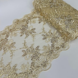 May include: A roll of gold-colored lace fabric with floral embroidery and sequins. The lace has a scalloped edge and is partially rolled, revealing the intricate design. The fabric is suitable for various craft and design projects.