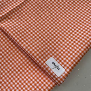 May include: Orange and white gingham fabric with a tag that reads "ORANGE". The fabric is folded, showing the pattern of small squares. The fabric is a warm orange color.