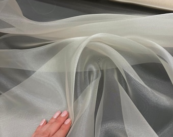 May include: Close-up of a sheer, off-white fabric with a flowing, translucent appearance. The fabric is draped in soft folds, creating a sense of movement and texture. The material appears lightweight and delicate, suitable for apparel or decorative projects.