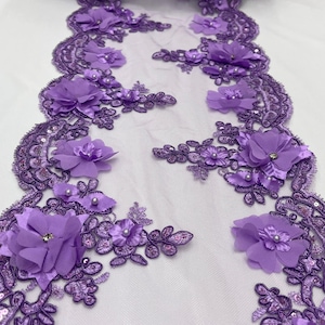 Puede incluir: Un adorno de encaje morado decorativo con diseños florales y de hojas. El adorno está adornado con lentejuelas, perlas de imitación y flores de tela 3D con centros de pedrería. El encaje está sobre un fondo de malla blanca.