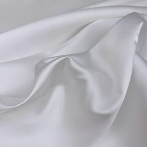 May include: A close-up shot of white fabric with soft folds and subtle shadows. The fabric appears to be smooth and possibly satin or silk, with a slight sheen. The image highlights the texture and flow of the material.
