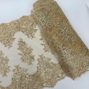 May include: A roll of gold lace fabric with intricate floral and scrollwork patterns. The lace is embellished with sequins, creating a shimmering effect. The fabric is displayed on a white surface.