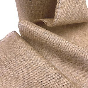 May include: Close-up of a roll of natural burlap fabric. The fabric is a light brown color with a coarse, woven texture. The edges are frayed, with a white thread visible along the border. The burlap is rolled and folded, showing its thickness and flexibility.