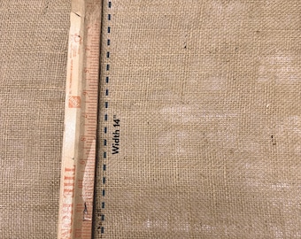 May include: A close-up of a piece of burlap fabric with a wooden ruler measuring its width. The burlap is a natural tan color, and the ruler indicates a width of 14 inches. The text "Width 14" is printed on the fabric.