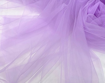 May include: A close-up of a soft, lavender tulle fabric. The sheer material is draped and layered, creating a delicate and airy texture. The fabric is a light purple color.