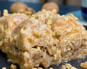 Puede incluir: Primer plano de una porción de baklava, un postre de hojaldre en capas con nueces y jarabe. La pasta dorada está cubierta con nueces picadas y un glaseado dulce. El postre está sobre un plato azul oscuro, con nueces esparcidas.