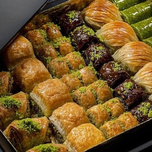 Może przedstawiać: Pudełko z różnymi ciastami baklava. Ciasta są złocistobrązowe, niektóre posypane zielonymi pistacjami, a inne ciemną polewą czekoladową. Pudełko jest czarne ze złotym wnętrzem, widoczna jest czarna wstążka.