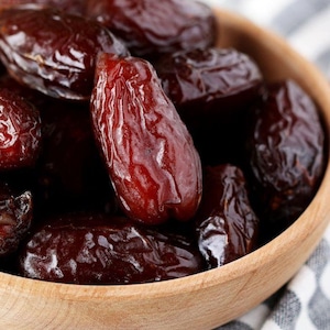Op de afbeelding: Een houten kom gevuld met donkerbruine dadels. De dadels hebben een glanzende, gerimpelde textuur en zijn in een close-up gerangschikt. De kom staat op een zwart-wit gestreepte doek.