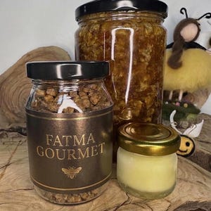 May include: Three glass jars of varying sizes with black and gold lids. The front jar features a dark brown label with the text FATMA GOURMET and a bee icon, filled with brown granules. A large jar contains amber honey, and a small jar holds cream.