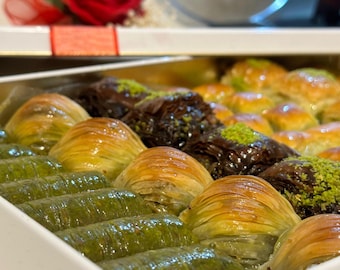 Puede incluir: Una bandeja rectangular blanca llena de varios tipos de baklava. Los pasteles son de color dorado, verde y chocolate, algunos cubiertos con pistachos. Una rosa roja y una cinta son visibles en el fondo.