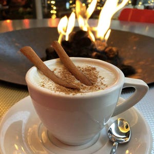 May include: A white ceramic cup of creamy beverage, garnished with cinnamon sticks and powder, sits on a saucer with a spoon. The background features a small fire pit with flames and blurred lights, creating a warm ambiance.
